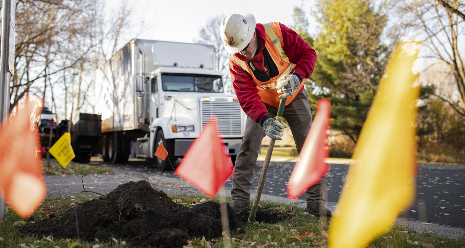 A coworker wearing safety gear digs a hole in the ground with a shovel.