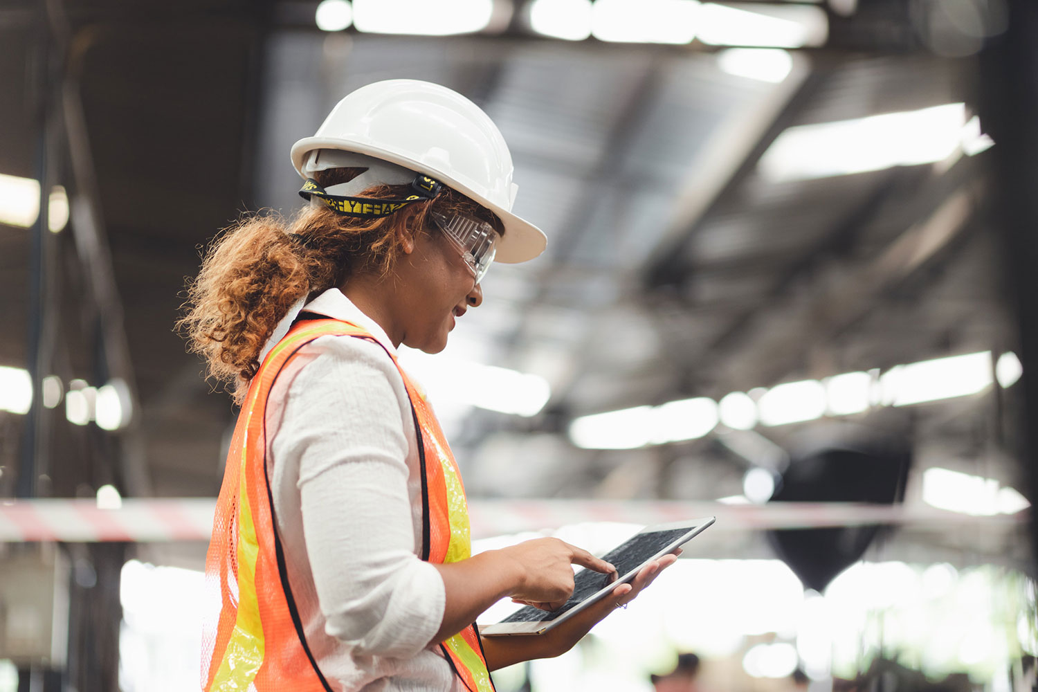 A woman wearing a white hardhat, safety goggles, and an orange safety vest works on her hand-held tablet computer.