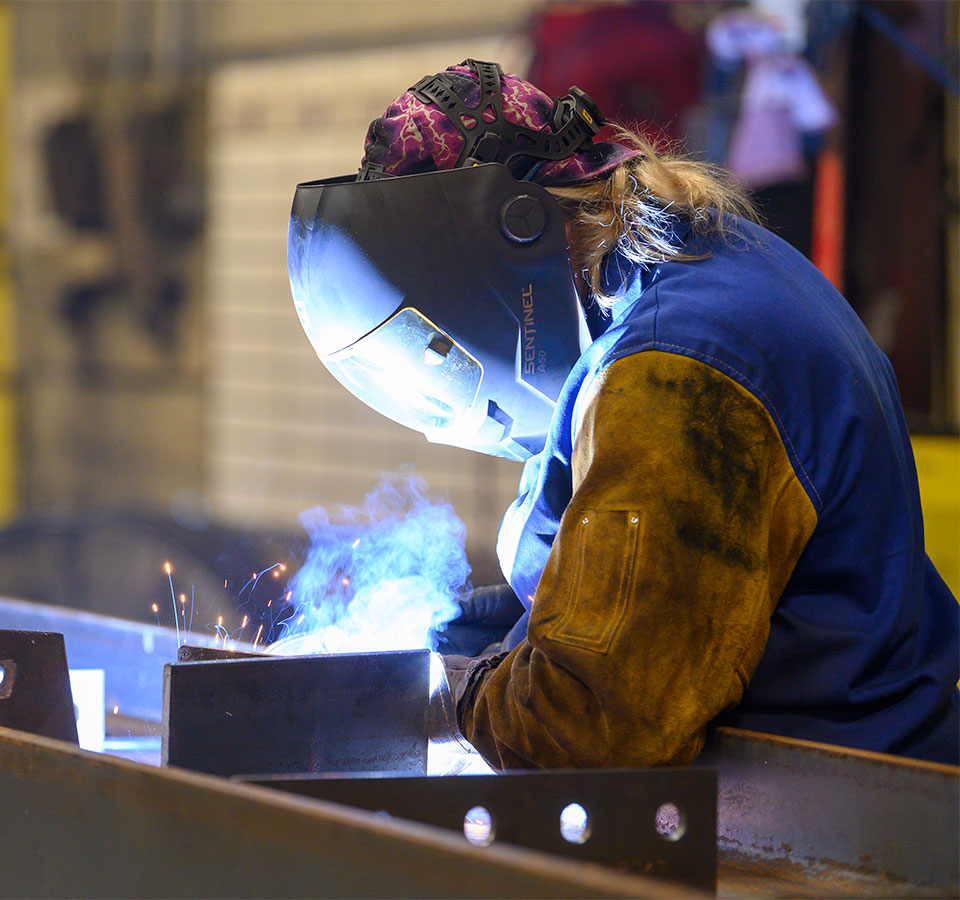 An employee wearing safety gear sands a metal pipe and sparks fly.